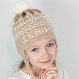 Puede incluir: Una persona joven lleva una gorra de punto beige con un pompón blanco y la palabra "CURIE" bordada en la parte delantera. La gorra tiene orejeras que se atan debajo de la barbilla.