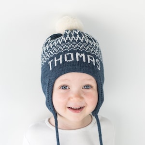 Puede incluir: Un gorro de punto azul oscuro con orejeras y cordones trenzados largos. El gorro presenta un patr&oacute;n geom&eacute;trico blanco en la parte superior y el nombre THOMAS tejido en letras blancas en el frente. Un pomp&oacute;n color crema corona el gorro.
