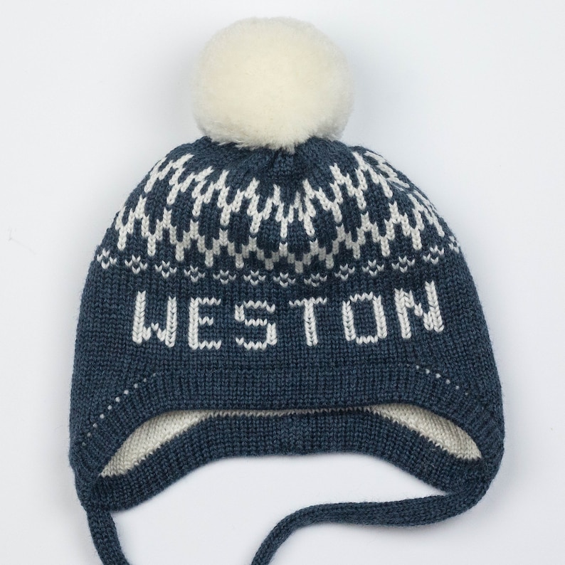 Puede incluir: Un gorro de punto azul oscuro con un pomp&oacute;n blanco en la parte superior, un patr&oacute;n geom&eacute;trico blanco alrededor de la corona y la palabra WESTON tejida en letras blancas en el frente. El gorro incluye orejeras y dos lazos largos de punto.