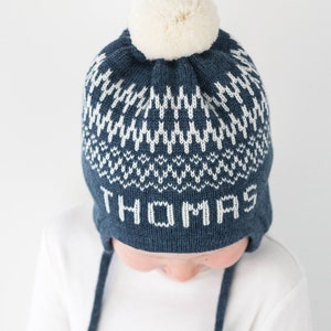 Puede incluir: Un gorro de punto azul marino con un patr&oacute;n geom&eacute;trico blanco y el nombre THOMAS tejido en letras blancas en la parte frontal. El gorro incluye orejeras con largos cordones trenzados azul marino y un pomp&oacute;n de lana color crema.
