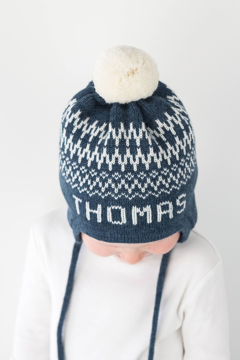 Puede incluir: Un gorro de punto azul marino con un patr&oacute;n geom&eacute;trico blanco y el nombre THOMAS tejido en letras blancas en la parte frontal. El gorro incluye orejeras con largos cordones trenzados azul marino y un pomp&oacute;n de lana color crema.