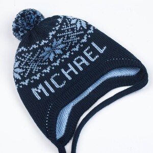 Puede incluir: Un gorro de punto azul marino con un pompón azul claro en la parte superior. El gorro presenta un patrón de copos de nieve azul claro alrededor de la corona y el nombre MICHAEL tejido en letras azul claro en el frente. Incluye orejeras.