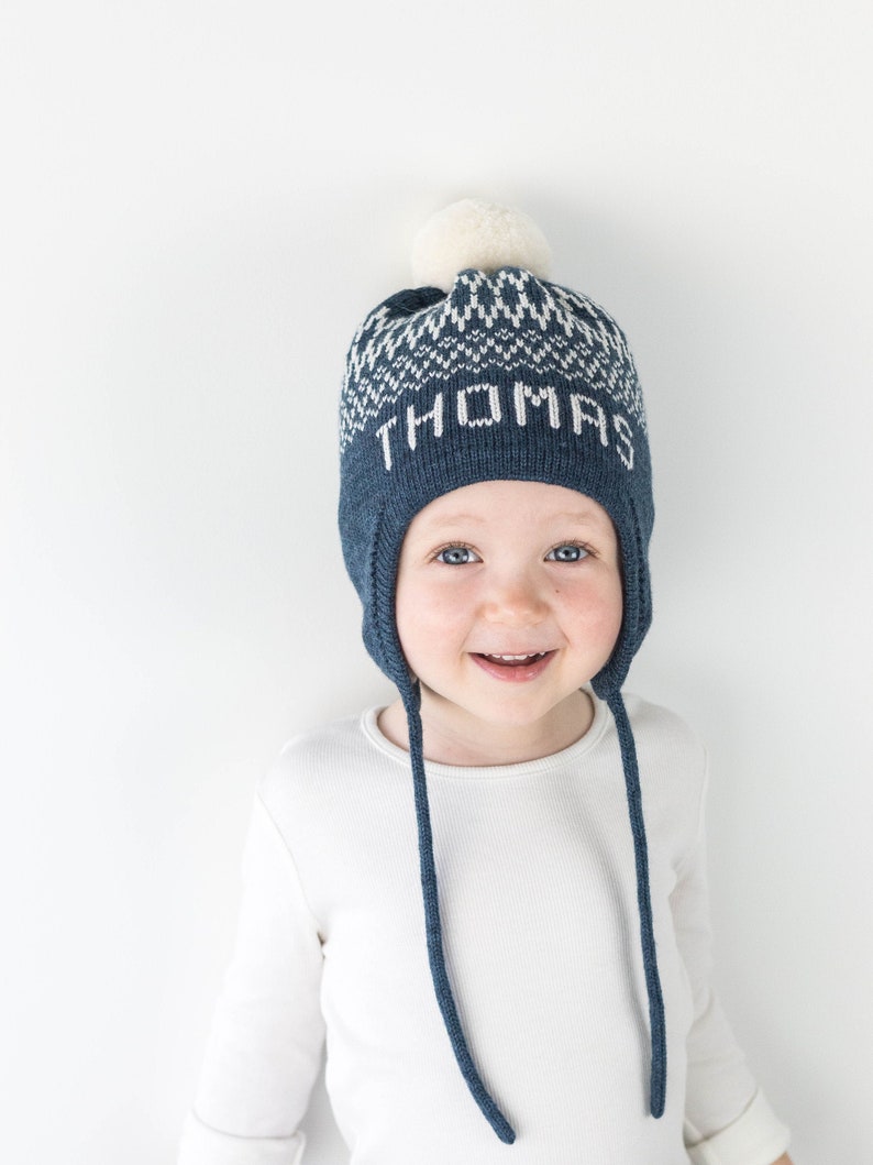 Puede incluir: Un gorro de punto azul oscuro con orejeras y cordones trenzados largos. El gorro presenta un patr&oacute;n geom&eacute;trico blanco en la parte superior y el nombre THOMAS tejido en letras blancas en el frente. Un pomp&oacute;n color crema corona el gorro.