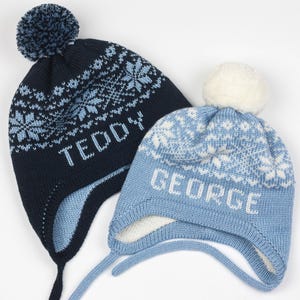 May include: Two blue and white knit winter hats with pom poms. The hats have a snowflake pattern and are personalized with the names "Teddy" and "George".
