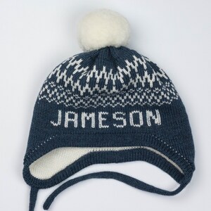 Puede incluir: Un gorro de punto azul oscuro con orejeras y un pomp&oacute;n blanco en la parte superior. El gorro presenta un patr&oacute;n geom&eacute;trico blanco alrededor de la corona y el nombre JAMESON tejido en letras blancas en el frente. Incluye dos cintas.