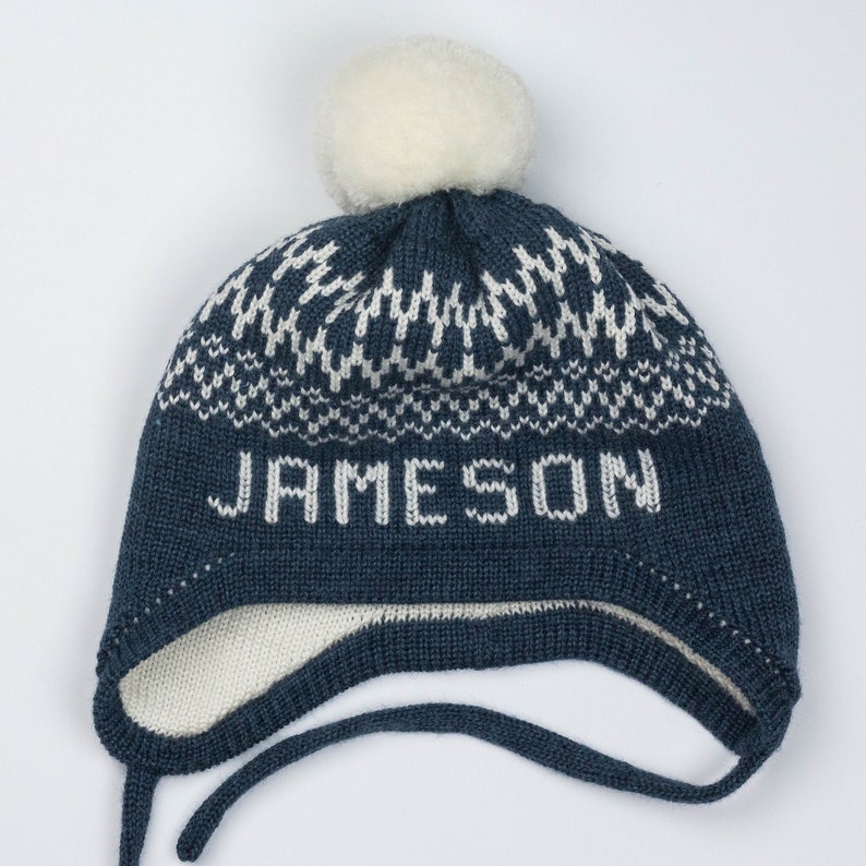 Puede incluir: Un gorro de punto azul oscuro con orejeras y un pomp&oacute;n blanco en la parte superior. El gorro presenta un patr&oacute;n geom&eacute;trico blanco alrededor de la corona y el nombre JAMESON tejido en letras blancas en el frente. Incluye dos cintas.