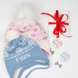 May include: Two knitted baby hats, one pink with hearts and one blue with a snowflake pattern. The blue hat has the name "Finn" embroidered on it. The hats are next to a gift box wrapped in white paper with a red ribbon and two cards with the text "hello baby" on them.