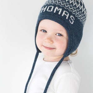 Puede incluir: Un gorro de punto azul oscuro con orejeras y lazos largos, con un patr&oacute;n geom&eacute;trico blanco y el nombre THOMAS tejido en blanco en la parte delantera. El gorro est&aacute; rematado con un pomp&oacute;n de color crema y tiene una textura acanalada.