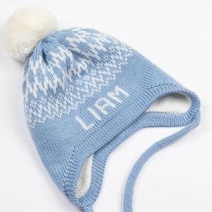 Puede incluir: Un gorro de punto azul claro con un pompón blanco en la parte superior, con un patrón geométrico blanco alrededor de la corona y el nombre LIAM bordado en letras blancas en el frente. El gorro incluye orejeras y dos lazos largos de punto.