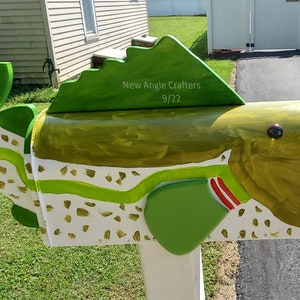 May include: A green, white, and gold painted fish-shaped mailbox with a red and white stripe on the side. The mailbox has a green fin on top and the text "New Angle Crafters 9/22" is written on the fin.