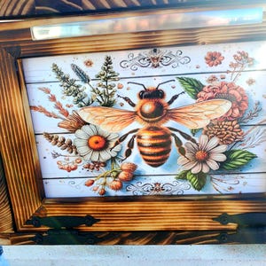 May include: Wooden box with a decorative bee and floral design. The artwork features a large bee with orange wings and a striped abdomen, surrounded by flowers and greenery. The box has a dark wood frame and hinges.