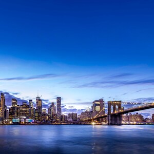 May include: A scenic cityscape at dusk, featuring a skyline of illuminated skyscrapers and the Brooklyn Bridge. The water reflects the city lights under a gradient blue sky with scattered clouds. The image captures the urban landscape.