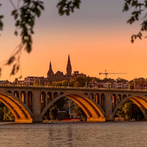May include: A view of a bridge over a river with a city skyline in the background. The sky is a vibrant orange and pink, creating a beautiful sunset scene.