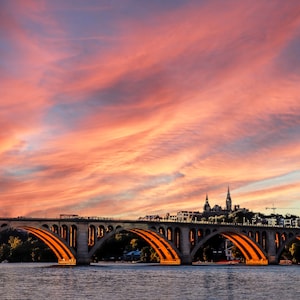 May include: A view of a bridge over a river with a city skyline in the background. The sky is a vibrant orange and pink with clouds.