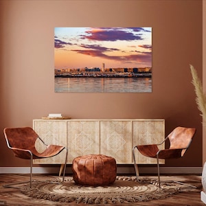K&ouml;nnte beinhalten: Ein Wohnzimmer-Interieur mit zwei braunen Ledersesseln, einem Schrank aus Holz mit kunstvollen Schnitzereien, einem braunen Lederhocker und einem beigefarbenen Teppich. Ein gro&szlig;es Bild einer Skyline einer Stadt bei Sonnenuntergang h&auml;ngt an der Wand.