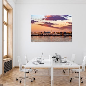 K&ouml;nnte beinhalten: Ein Panoramafoto einer Skyline bei Sonnenuntergang. Der Himmel ist ein lebendiges Gemisch aus rosa, lila und orangefarbenen Farbt&ouml;nen mit flauschigen Wolken. Die Stadt ist vor dem farbenfrohen Himmel silhouettiert, mit einem hohen Geb&auml;ude in der Mitte.