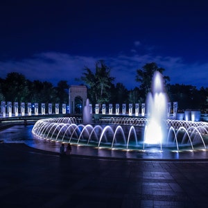 May include: A night view of the World War II Memorial in Washington, D.C. The memorial is lit up with blue lights, and there are fountains in the reflecting pool. The memorial is made of white marble and features a large archway.