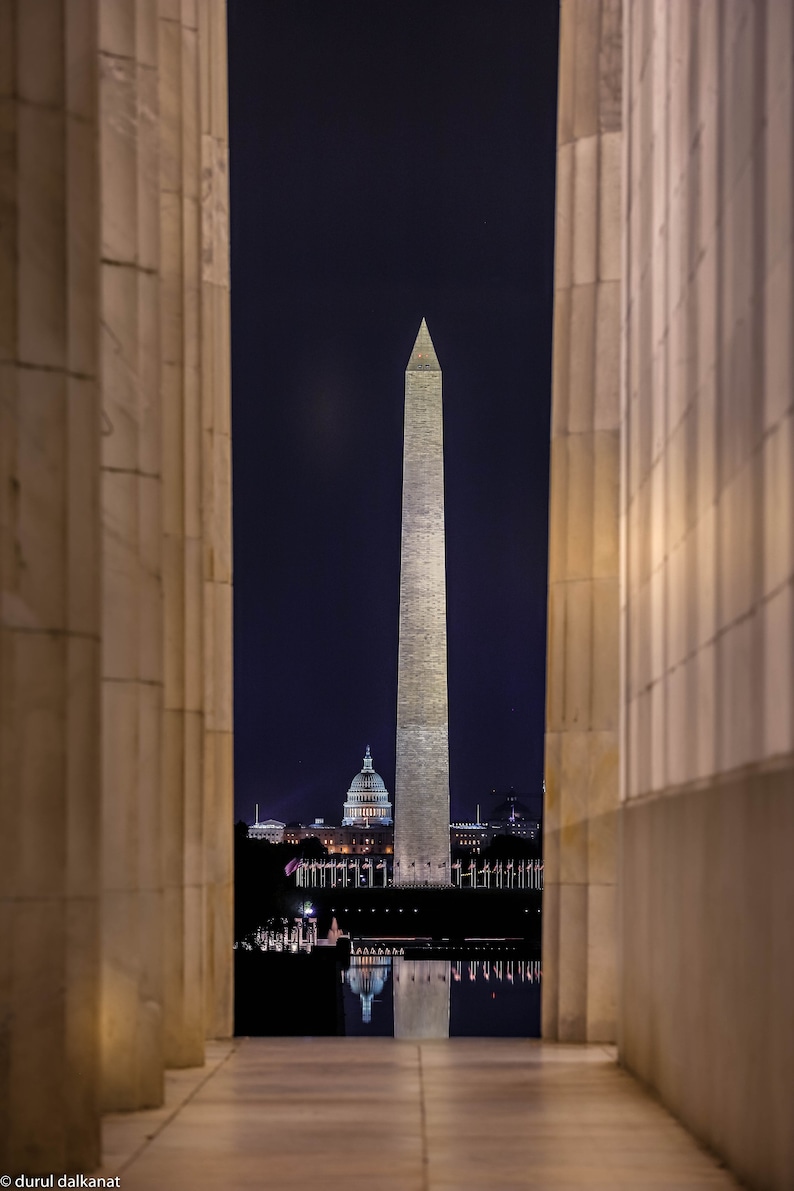 May include: A view of the Washington Monument and the Capitol Building in Washington, D.C. at night. The buildings are illuminated and reflected in the water. The view is framed by two columns.