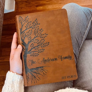 May include: A tan leather-like journal featuring a dark brown laser-engraved tree design on the front cover. The text reads The Anderson Family and EST 09.30.23. The cover has a smooth texture and a rectangular shape with rounded corners.