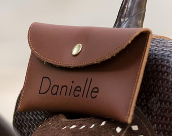 Może przedstawiać: Brązowa skórzana saszetka z klapką i złotym zapięciem na zatrzask. Na przodzie wytłoczono imię "Danielle". Saszetka spoczywa na siodle.