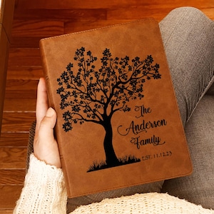 May include: A brown rectangular leather-like journal featuring a black laser-engraved design of a tree with foliage. The text "The Anderson Family" and "EST. 11.12.23" is engraved in black on the right side of the cover, centered below the tree branches.
