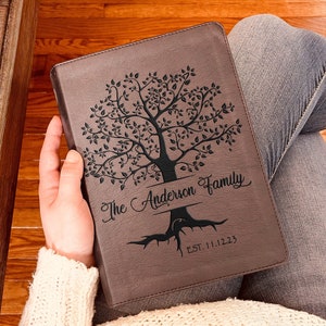 May include: A brown rectangular leather-like book cover featuring a laser-engraved tree design. The text "The Anderson Family" is centered on the trunk, with "EST. 11.12.23" engraved below the roots. The cover has stitched edges and a smooth finish.