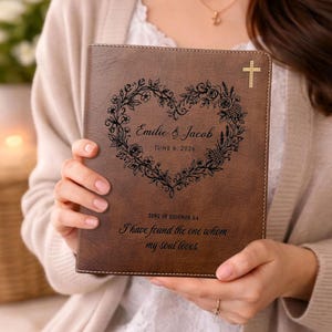 May include: A brown leather-effect journal featuring a laser-engraved floral heart design, a small cross in the top right corner, and the text Emilie & Jacob, June 6, 2026, and Song of Solomon 3:4, I have found the one whom my soul loves.