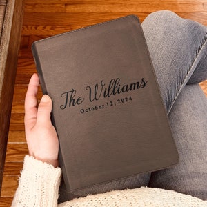 May include: A dark gray rectangular leather-like journal with a smooth, matte finish. The front cover features the engraved text "The Williams" in a cursive script font, followed by the printed date "October 12, 2024" in a simple sans-serif font.