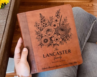May include: A tan leather-like binder featuring a laser-engraved floral bouquet at the top. The text reads The LANCASTER family, October 12, 2024, and the names Robert, Shannon, Andrei, Emma, Riley, and Alex are engraved at the bottom.