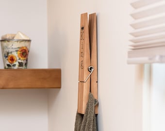 May include: A large wooden clothespin-shaped towel holder mounted on a wall. The word "KING" is vertically etched on the holder. A gray towel is clipped to the holder. A small bucket with a sunflower design sits on a shelf in the background.