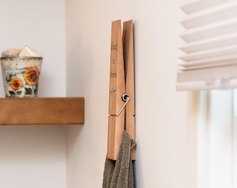 May include: A large wooden clothespin-shaped wall hook with the words "Hang in there" etched on the side. A gray towel hangs from the hook. The hook is mounted on a light-colored wall.