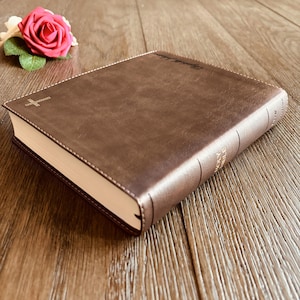 May include: A brown leather-bound Holy Bible with visible stitching along the edges, a small gold cross emblem on the front cover, and the text Holy Bible embossed on the spine. The book is positioned next to a single pink rose with green leaves.