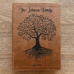 Pode incluir: Um diário em couro sintético castanho-claro com um design de árvore genealógica gravado a laser em preto. O texto diz The Johnson Family no topo e Est. 2023 na base. Os nomes Dave, Becky, Claire, Marie, Louise e James estão gravados.