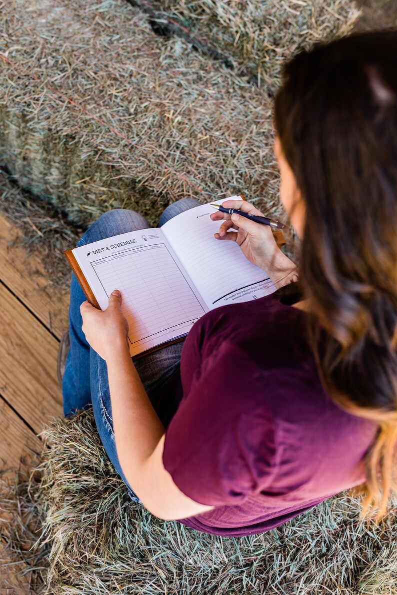 May include: A person is writing in a Diet & Schedule journal with a brown leather cover. The journal is open to a blank page with lines and the text "Diet & Schedule". The person is wearing a burgundy shirt and blue jeans.