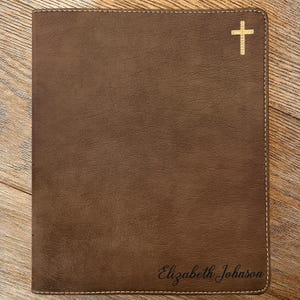 May include: A brown rectangular leather cover featuring a textured surface and light-colored stitching along the edges. A gold-colored cross icon is positioned in the top right corner, and the name Elizabeth Johnson is engraved in the bottom right.
