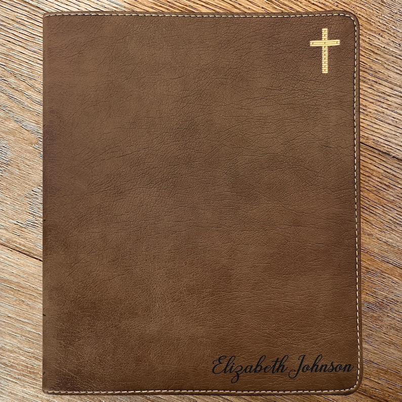 May include: A brown rectangular leather cover featuring a textured surface and light-colored stitching along the edges. A gold-colored cross icon is positioned in the top right corner, and the name Elizabeth Johnson is engraved in the bottom right.