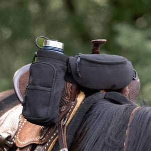 May include: Black saddle accessories, including a water bottle holder and a zippered pouch, attached to a brown leather saddle. The water bottle holder has a small pocket and a silver-coloured lid. The background is blurred green.