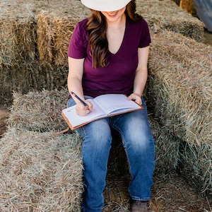 May include: A person wearing a white cowboy hat with a patterned band and a dark purple V-neck shirt, writing in a brown leather journal. The person is sitting on a stack of hay bales, wearing blue jeans and brown cowboy boots.
