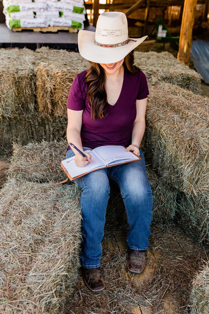 May include: A person wearing a white cowboy hat with a patterned band and a dark purple V-neck shirt, writing in a brown leather journal. The person is sitting on a stack of hay bales, wearing blue jeans and brown cowboy boots.