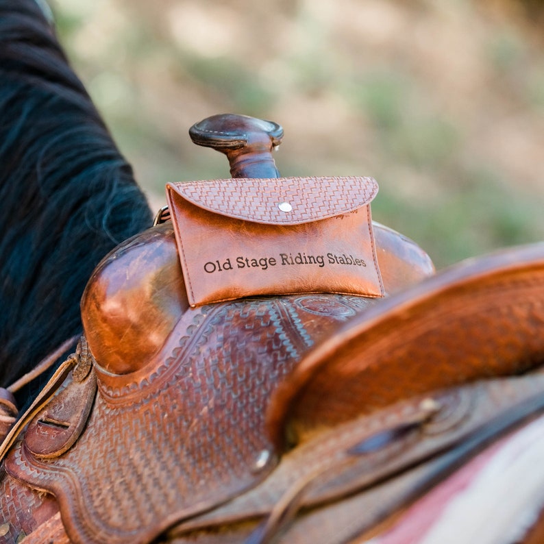 K&ouml;nnte beinhalten: Ein brauner Ledersattel mit einer kleinen braunen Ledertasche, die an der Vorderseite befestigt ist. Die Tasche tr&auml;gt den gepr&auml;gten Text "Old Stage Riding Stables".