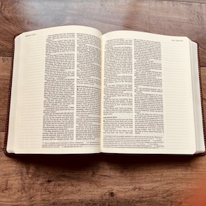 May include: An open book with two pages of printed text in columns. The pages contain the biblical text of Ruth chapters 3 and 4, featuring black ink on off-white paper. The book has a dark brown cover and is displayed on a wooden surface.