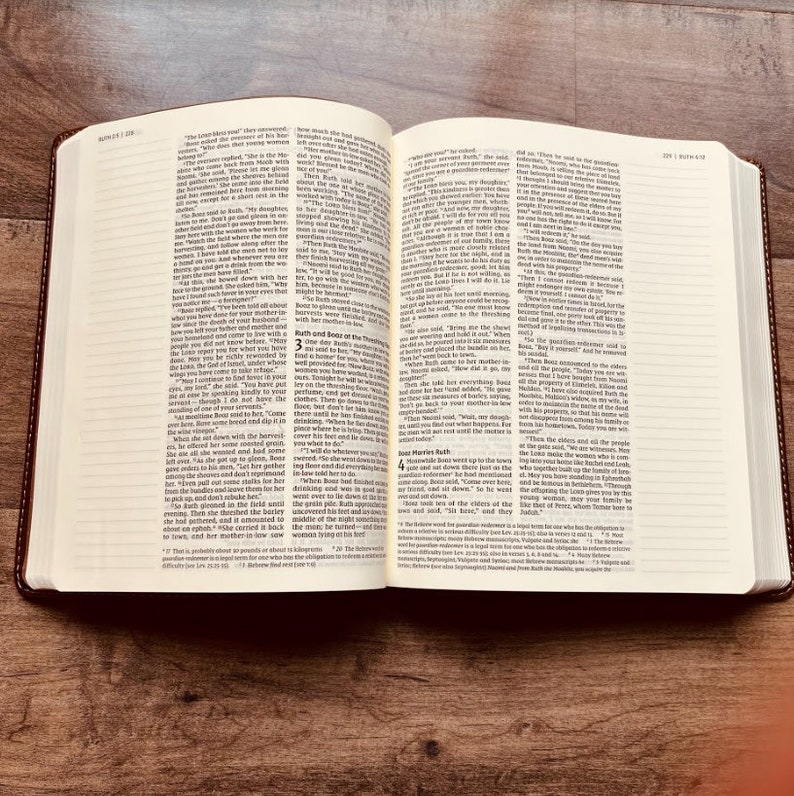 May include: An open book with two pages of printed text in columns. The pages contain the biblical text of Ruth chapters 3 and 4, featuring black ink on off-white paper. The book has a dark brown cover and is displayed on a wooden surface.