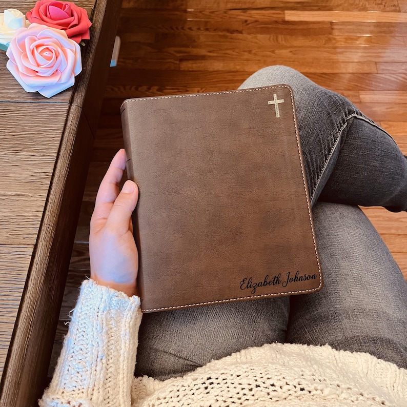 May include: A brown leather-like book cover featuring a small white cross icon at the top right corner and the name Elizabeth Johnson engraved in a black script font at the bottom right corner, with visible white stitching along the edges.