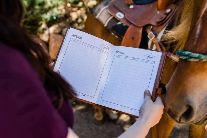 Puede incluir: Un cuaderno de cuero marr&oacute;n con las palabras "ARENAS" y "TRAILS" impresas en la portada. El cuaderno est&aacute; abierto en dos p&aacute;ginas con secciones cuadriculadas para escribir. Una persona est&aacute; sosteniendo el cuaderno frente a un caballo marr&oacute;n.