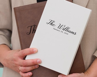 May include: A white and brown leather-bound journal with the text "The Williams January 24, 2026" on the front cover. The journal is held in front of a brown leather journal.