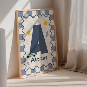 Op de afbeelding: Een ingelijste kunstprint met de letter 'A' in blauw en de naam 'Atticus' eronder. De achtergrond heeft een lichtblauw en crèmekleurig dambordpatroon, met een ster, een bliksemschicht en een lachende zon. De lijst is van licht hout.