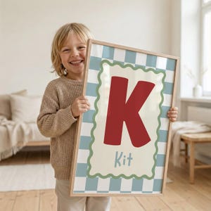 Puede incluir: Un estampado enmarcado con la letra "K" roja sobre un fondo crema, con la palabra "Kit" debajo. El estampado tiene un borde a cuadros azul claro y blanco y un borde festoneado verde. El marco es de madera clara.