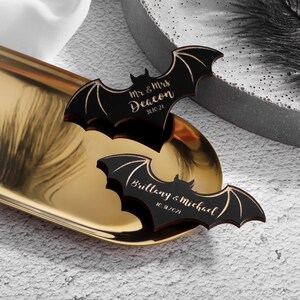 May include: Two black wooden bat-shaped wedding favours with gold lettering. The top bat reads "Mr. & Mrs. Deacon 3.10.21". The bottom bat reads "Brittany & Michael 10.2.21".