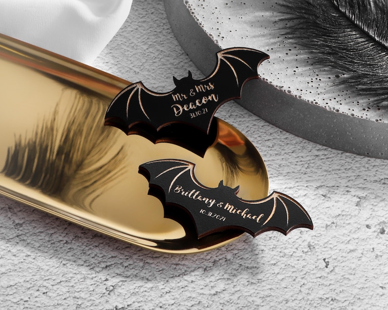 May include: Two black wooden bat-shaped wedding favours with gold lettering. The top bat reads "Mr. & Mrs. Deacon 3.10.21". The bottom bat reads "Brittany & Michael 10.2.21".
