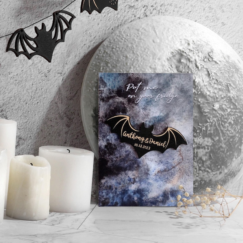 May include: A black wooden bat with gold lettering that says "Put me on your fridge" and "Anthony & Daniel 10.13.2023" on a dark blue and grey watercolour background.